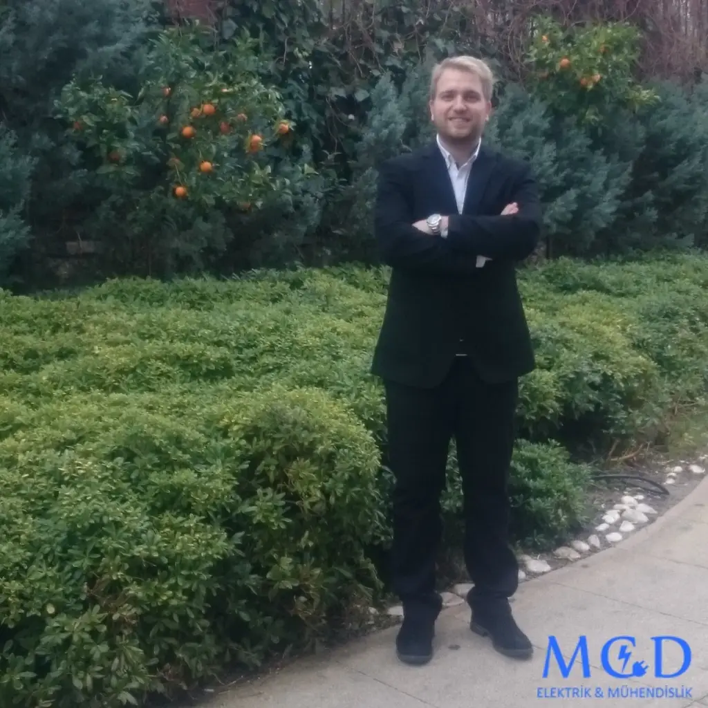 MCD Elektrik Mühendislik logosunun yer aldığı, Hakkımızda sayfasında Mustafa Caner Demir’in takım elbiseyle bir bahçede kollarını çapraz yapmış şekilde durduğu fotoğraf; arka planda yeşil çalılar ve portakal ağaçları görünüyor.