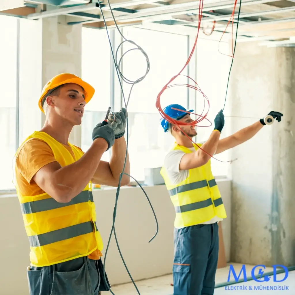 MCD Elektrik Mühendislik logosunun yer aldığı, iki teknisyenin bir inşaat alanında elektrik tesisatı kablolarını düzenlediği, güvenlik yelekleri ve baretleriyle çalıştığı bir görsel.