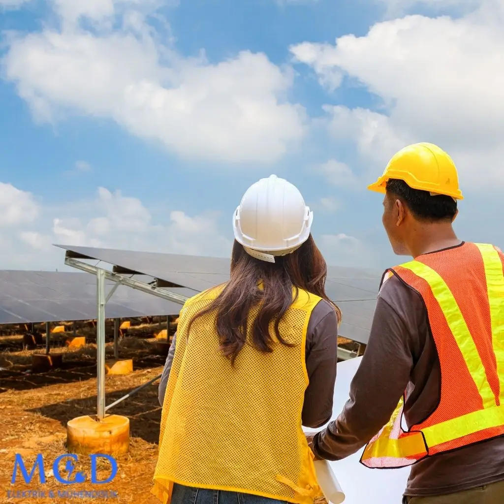 MCD Elektrik Mühendislik logosunun yer aldığı, güneş enerjisi panellerinin önünde planları inceleyen, güvenlik yelekleri ve baretleriyle iki uzman ekip üyesinin çalıştığı bir görsel; arka planda açık bir gökyüzü ve doğal bir alan görünüyor.